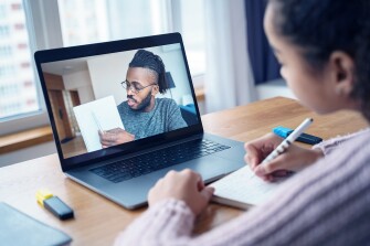 View on laptop of a Black male teacher with a young student sitting at a desk.