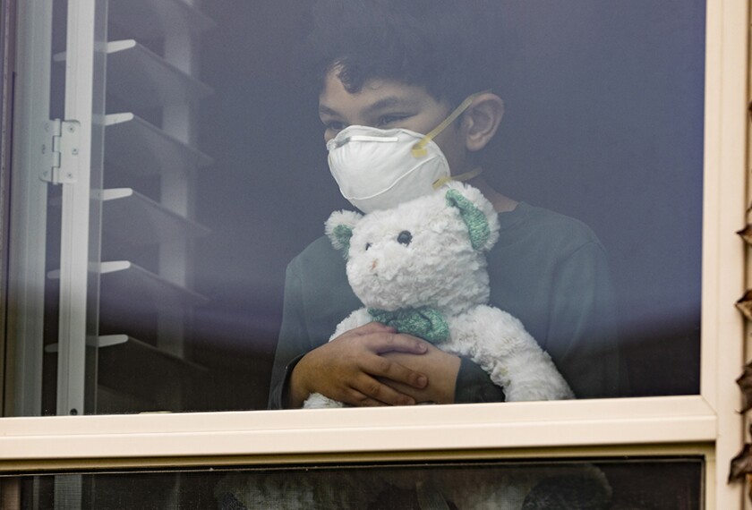 Young boy wearing a mask shown sheltering at home looking out a window with a stuffed animal.
