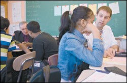 Roger Brown, left, of Cambridge Education, gathers information from students at the International High School at Prospect Heights in Brooklyn as mathematics teacher Todd Grunow, right, works with another student. The British company is helping the New York City schools pilot-test part of a new accountability system.