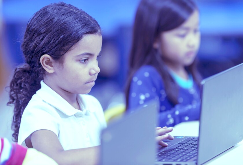 A young, culturally diverse elementary student is typing on a laptop as she sits at her desk. There are diverse kids blurred on either side of her working on laptops.