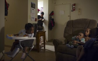 Kourtney Mills peers out from the kitchen to check on her children Emily Bakker, 20 months, left, Blake Bakker, 11 months, center, and Nathan Mills, 8, right, while eating her breakfast. Mills says living in public housing has provided stability for her children, allowing them to take the same walk to school every morning and return to the same bed every night.