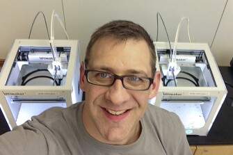 Zachary Carrico, an engineering teacher at a career and technical education center in Johnstown, N.Y., stands in front of two of his center’s 3-D printers, which are making face shields for medical workers.