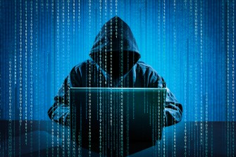 Silhouette of a hacker in a hoodie using laptop with binary code overlay.